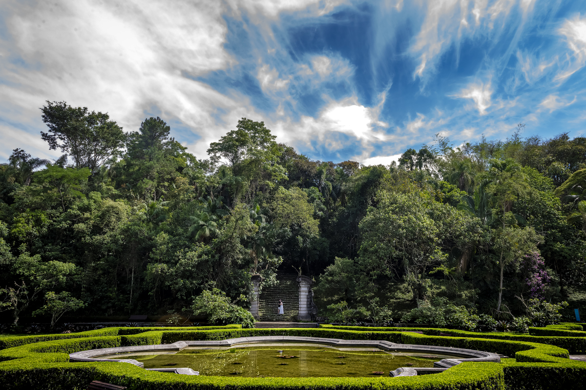 Plano panorâmico do Jardim Botânico de São Paulo com céu dramático, escadaria histórica e espelho d'água, casal de noivos ao fundo.
