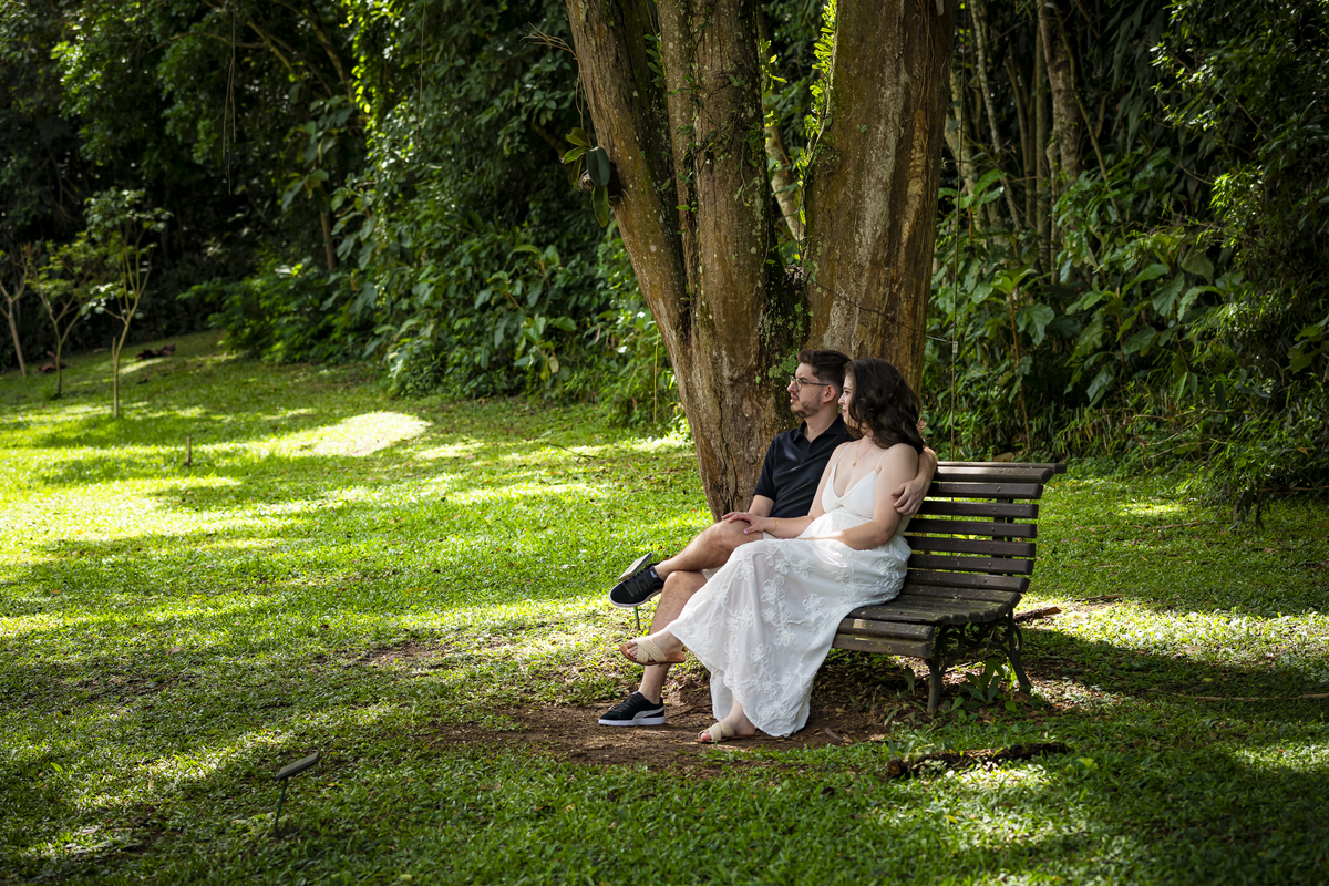 Casal de noivos sentado em banco de madeira sob árvore frondosa, em momento contemplativo no Jardim Botânico de São Paulo.