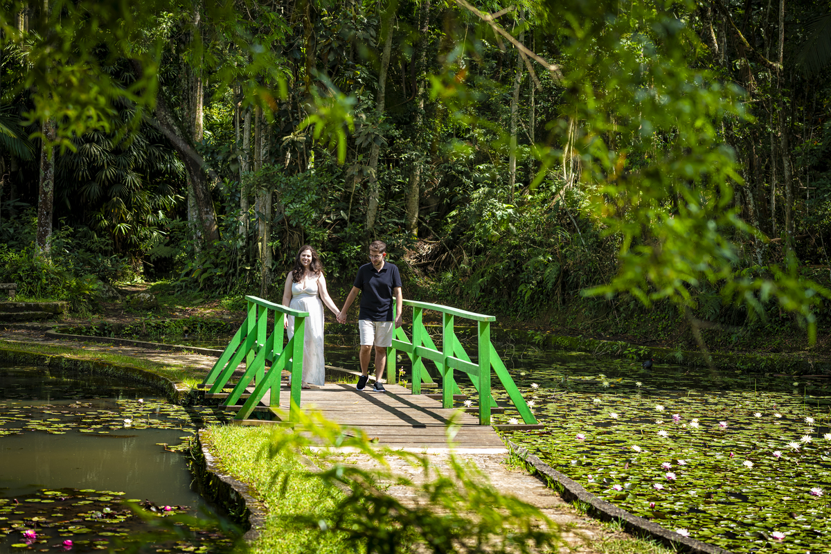 Casal de noivos atravessa pequena ponte verde sobre lago de vitórias-régias floridas no Jardim Botânico de São Paulo.