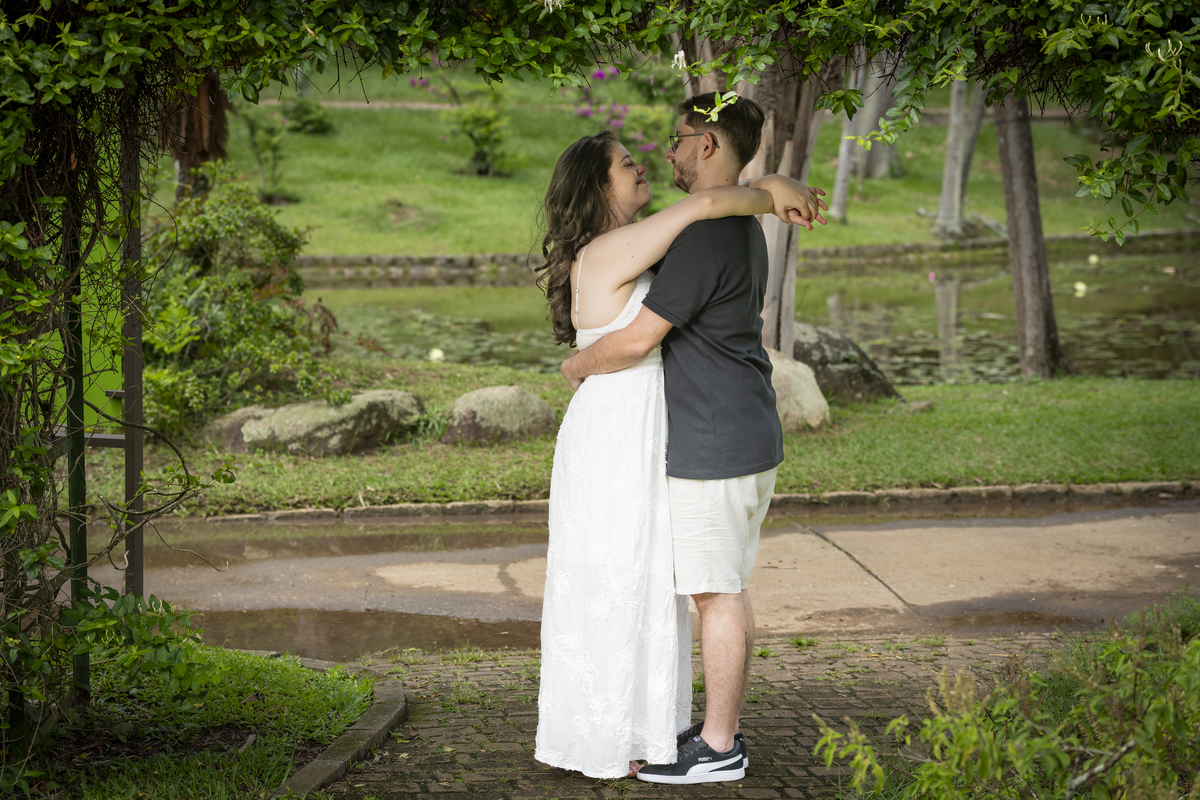 Abraço do casal de noivos sob arco de folhagem florida, com lago ao fundo, no Jardim Botânico de São Paulo.
