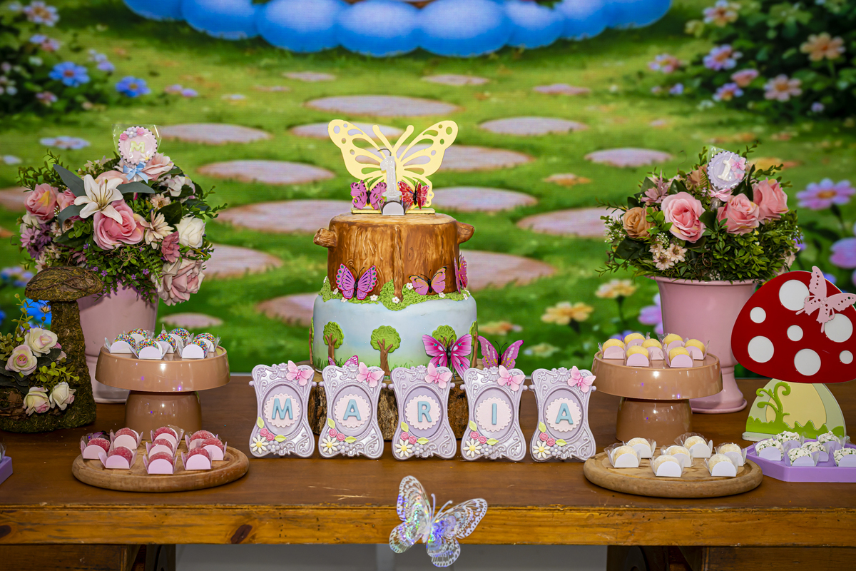 Detalhe do bolo de aniversário com tema de tronco e borboletas, cercado por docinhos personalizados.