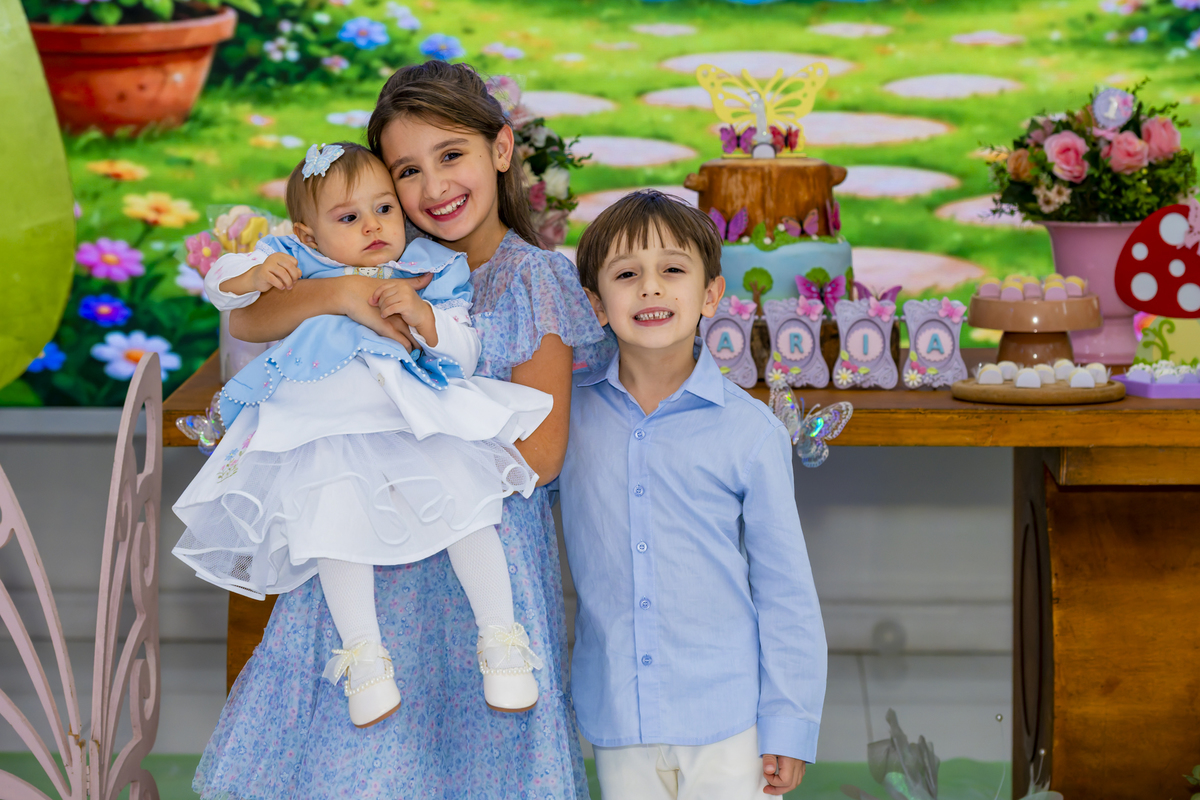 Os irmãos sorridentes segurando a aniversariante no colo, diante da mesa de doces decorada.