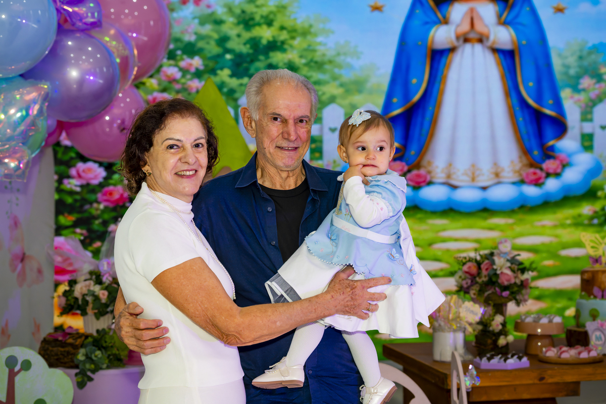 Avós sorridentes segurando a aniversariante em frente ao painel temático de jardim encantado.