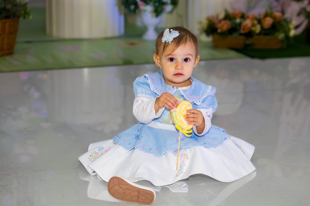 Bebê sentada no chão espelhado segura docinho durante festa de aniversário em Alphaville. O vestido azul e branco combina com a decoração floral do salão Lá Vem o Sol - Lua.