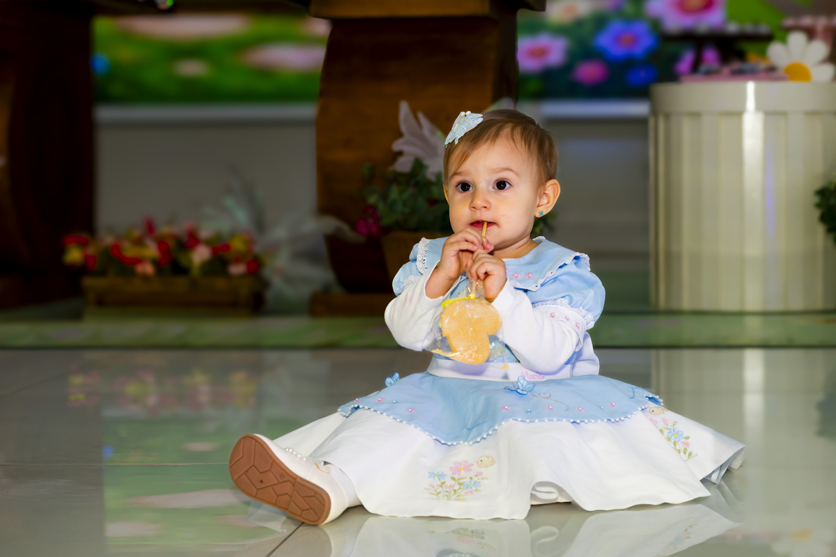 Aniversariante saboreia doce sentada no piso brilhante da festa infantil, com cenário lúdico ao fundo no buffet Lua, em Alphaville. Detalhe do vestido azul reforça o tema clássico da comemoração de um aninho.