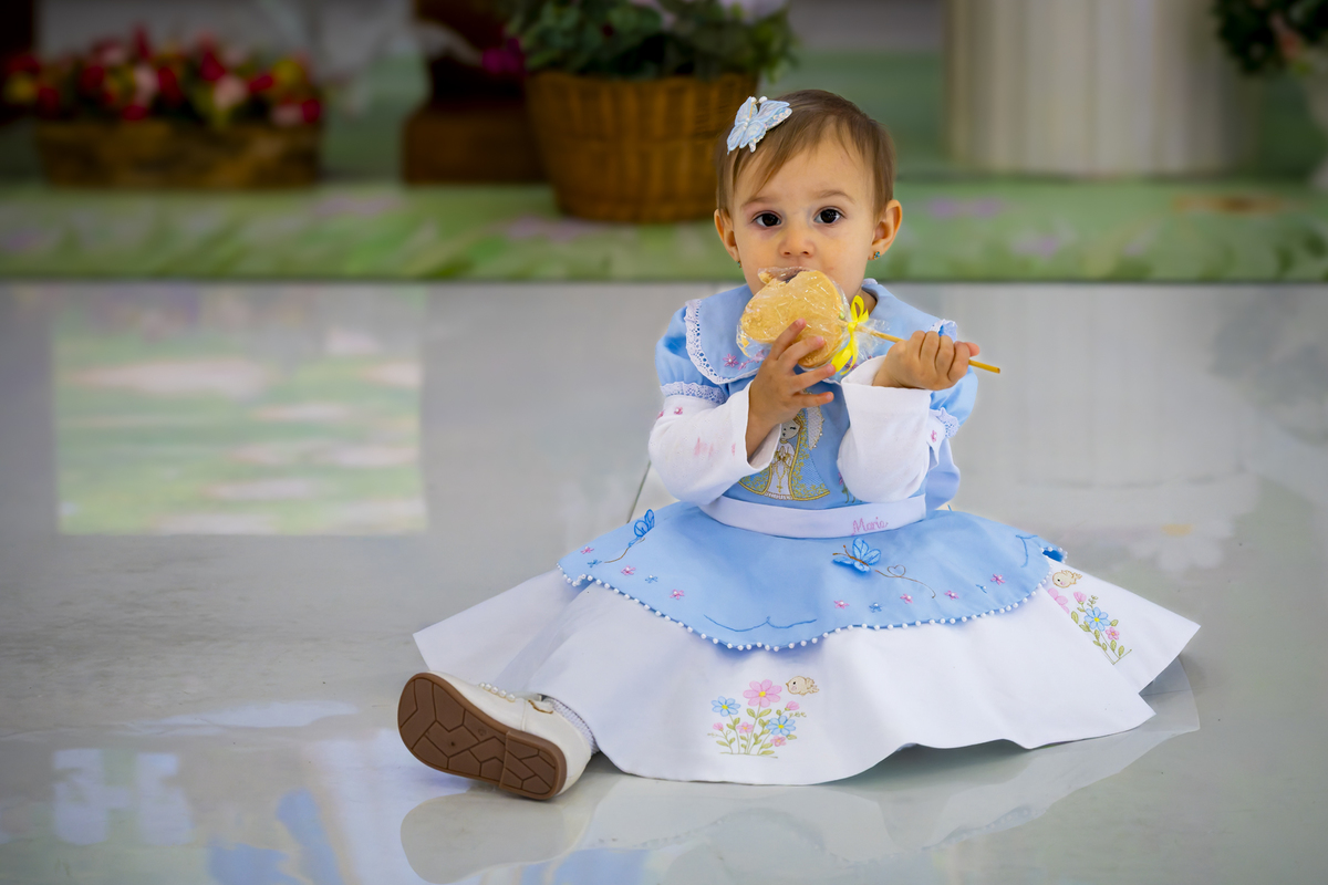 Bebê concentrada mordendo doce durante sessão fotográfica de aniversário no espaço Lá Vem o Sol, em Alphaville. Vestido azul bordado e sapatinho branco reforçam a fofura do registro de um aninho.