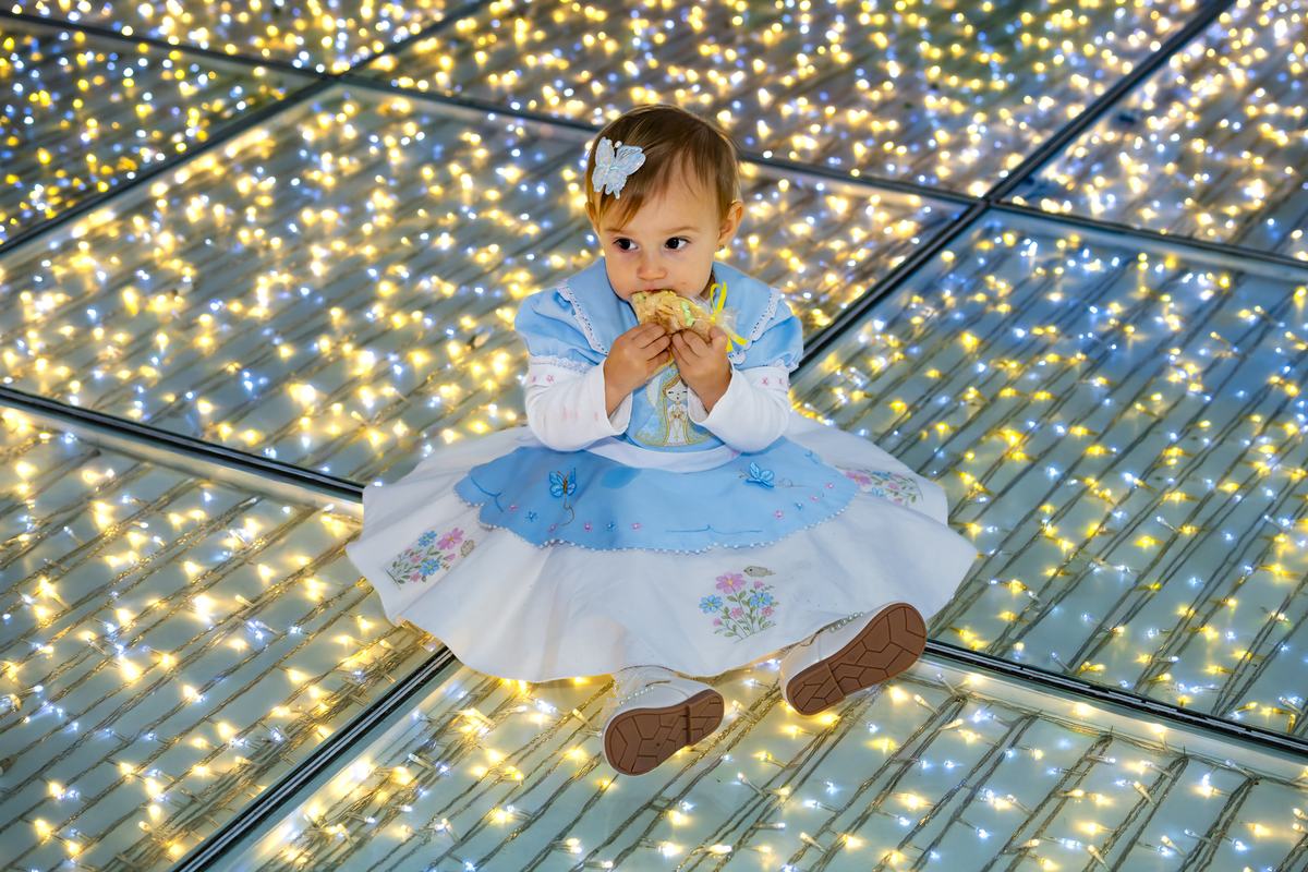 Aniversariante sentada sobre piso iluminado com luzinhas douradas, saboreando docinho durante festa em Alphaville. O efeito de luzes cria um cenário mágico para o ensaio de um aninho.