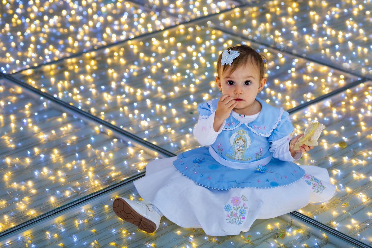 Bebê leva a mão à boca em pose espontânea sobre piso de luzes na festa infantil do espaço Lua, em Alphaville. O vestido azul claro e a presilha de borboleta destacam o estilo delicado da produção.