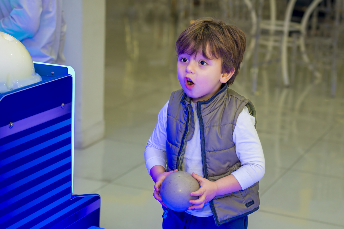 Menino brinca com bola em brinquedo interativo iluminado durante festa de aniversário no espaço Lá Vem o Sol, em Alphaville. Expressão de surpresa registra a diversão típica das comemorações infantis no local.