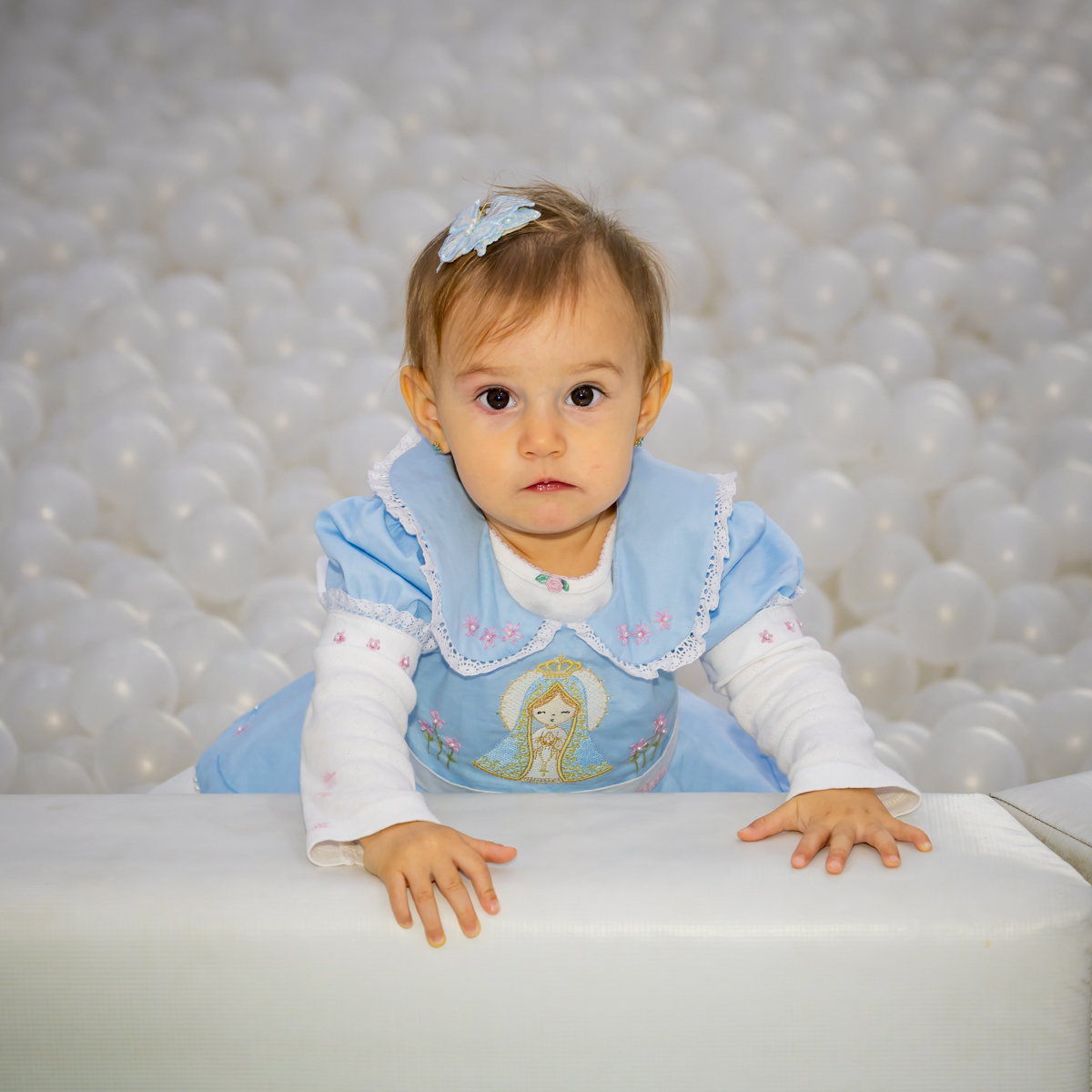 Aniversariante em vestido azul aparece por trás de piscina de bolinhas brancas durante festa em Alphaville. O olhar curioso e as mãos apoiadas compõem um momento espontâneo da celebração.
