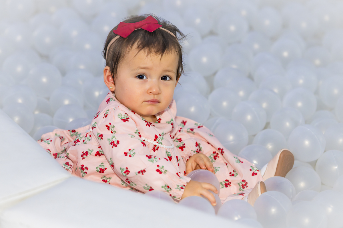 Convidada em vestido floral rosa observa a câmera sentada na piscina de bolinhas durante aniversário infantil em Alphaville. Laço vermelho no cabelo complementa o look da pequena convidada.