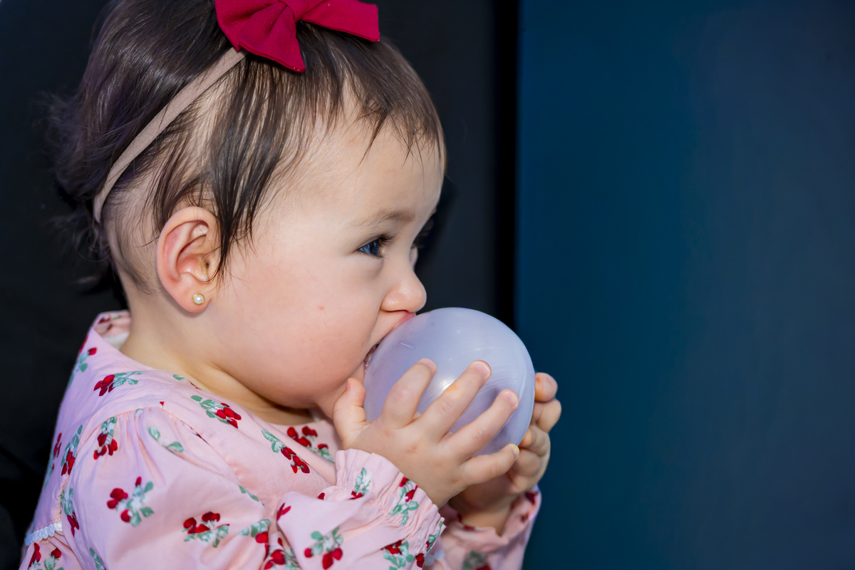 Criança pequena morde bolinha de brinquedo em registro descontraído na festa de aniversário no espaço Lua, em Alphaville. O laço vermelho e o vestido floral trazem charme à cena espontânea.