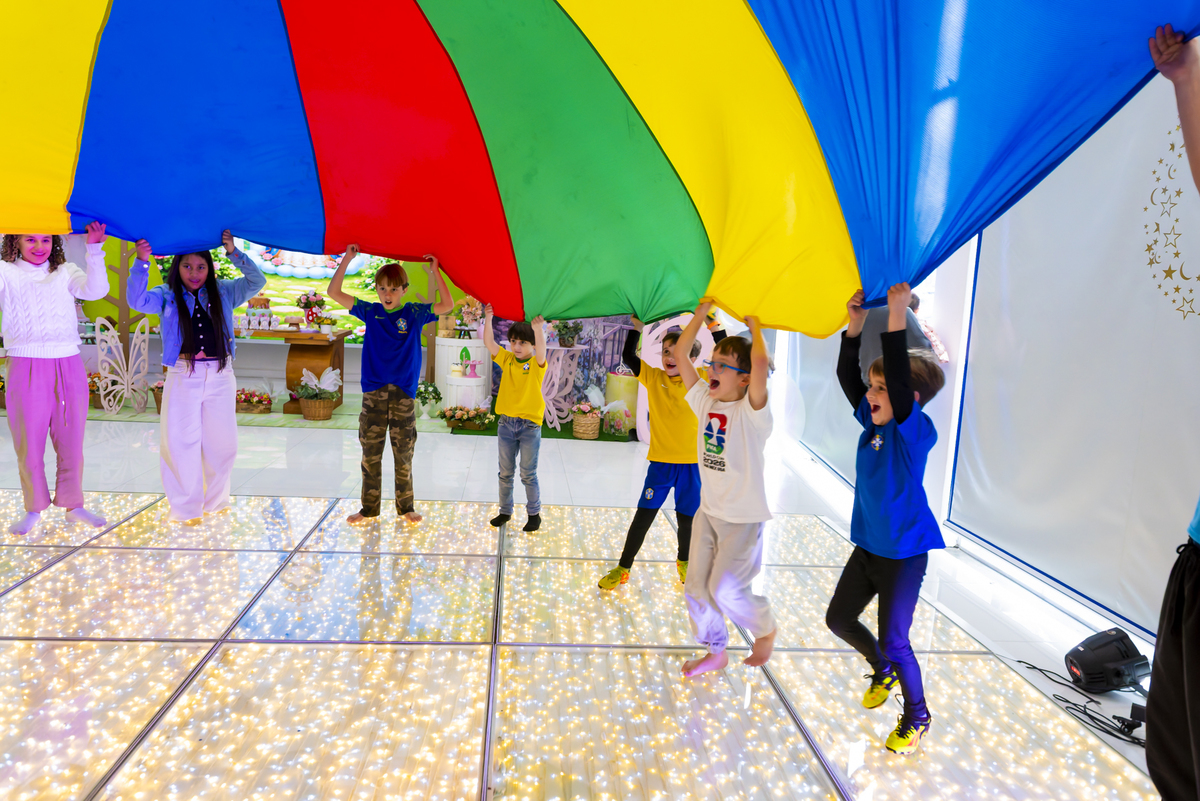 Crianças erguem paraquedas multicolorido em brincadeira animada durante aniversário no espaço kids em Alphaville.