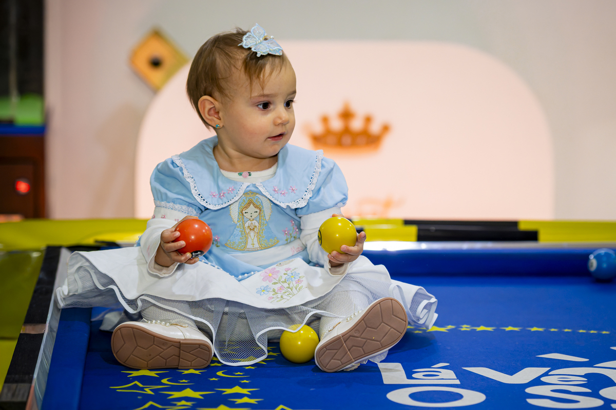 Bebê vestida de azul segura bolinhas coloridas sentada sobre mesa de sinuca personalizada da festa.