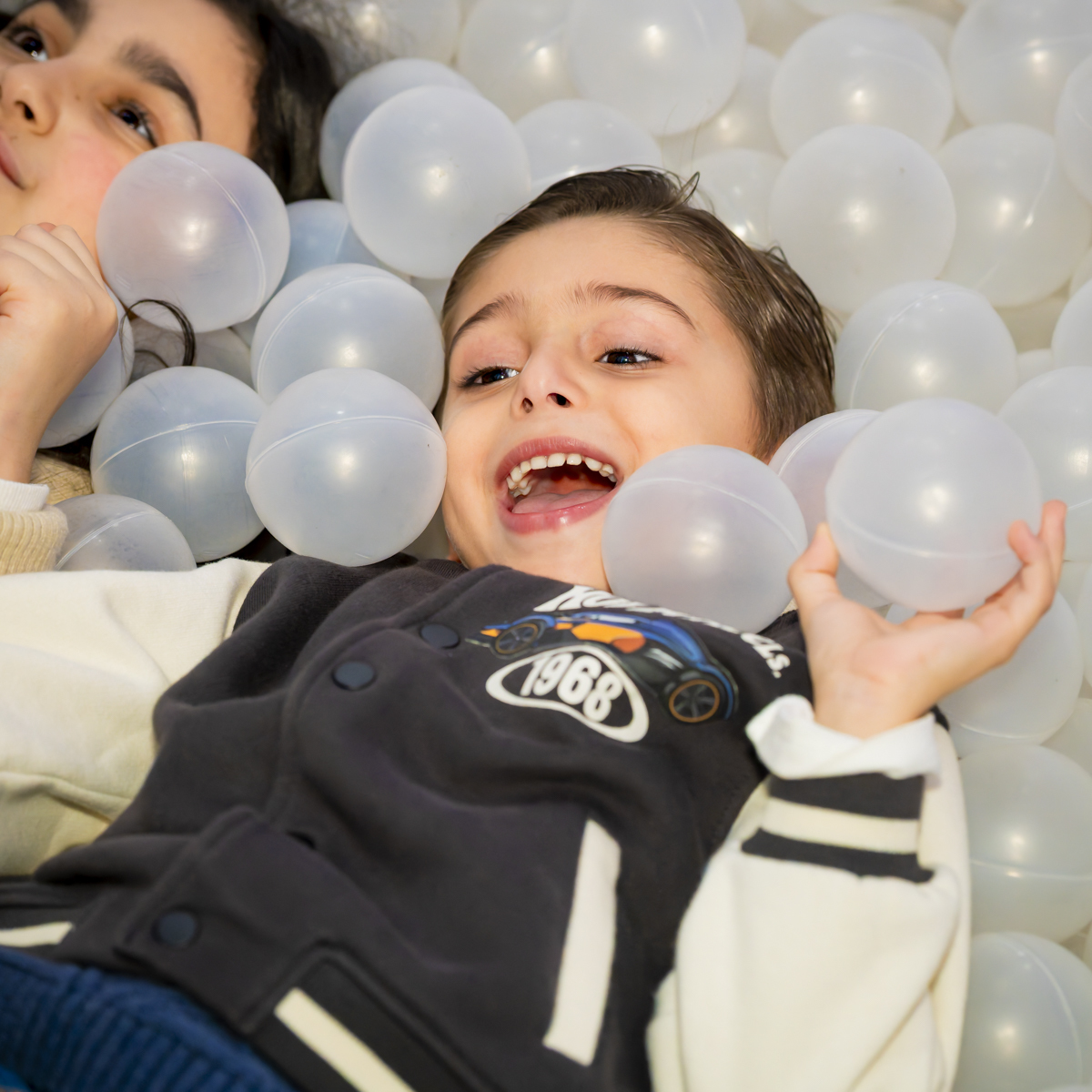 Menino sorridente brinca na piscina de bolinhas brancas durante festa de aniversário no espaço Lá Vem o Sol - Lua, em Alphaville. Momento espontâneo de diversão infantil registrado com luz natural.