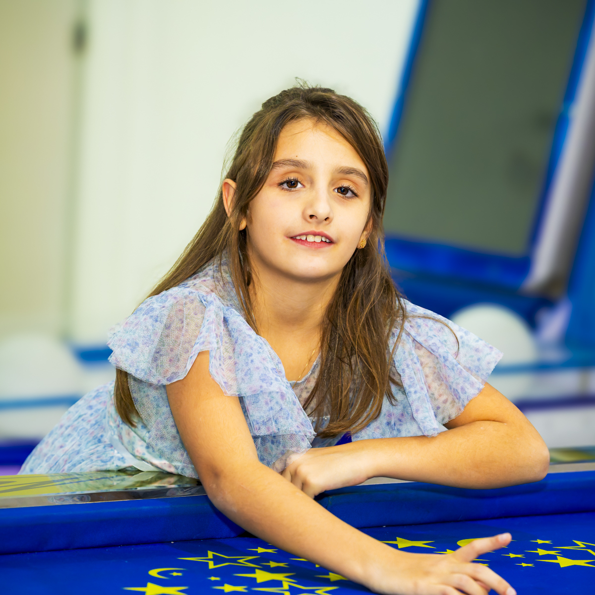 Aniversariante posa apoiada no air hockey com vestido floral azul, no salão de festas infantil Lá Vem o Sol - Lua, Alphaville. Retrato descontraído com fundo desfocado do espaço de jogos.