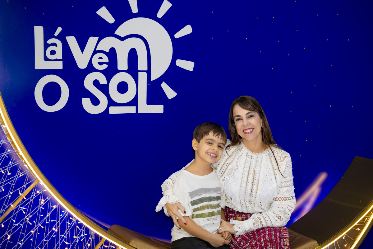 Convidado e sua mãe sorriem abraçados junto ao painel decorativo 'Lá Vem o Sol', destaque da ambientação temática da festa em Alphaville.