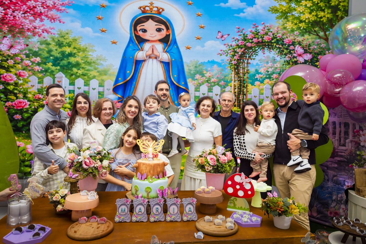 Família reunida posa sorridente ao lado da mesa do bolo, com cenário lúdico de jardim e imagem religiosa infantil, durante celebração de aniversário no buffet Lá Vem o Sol - Lua.