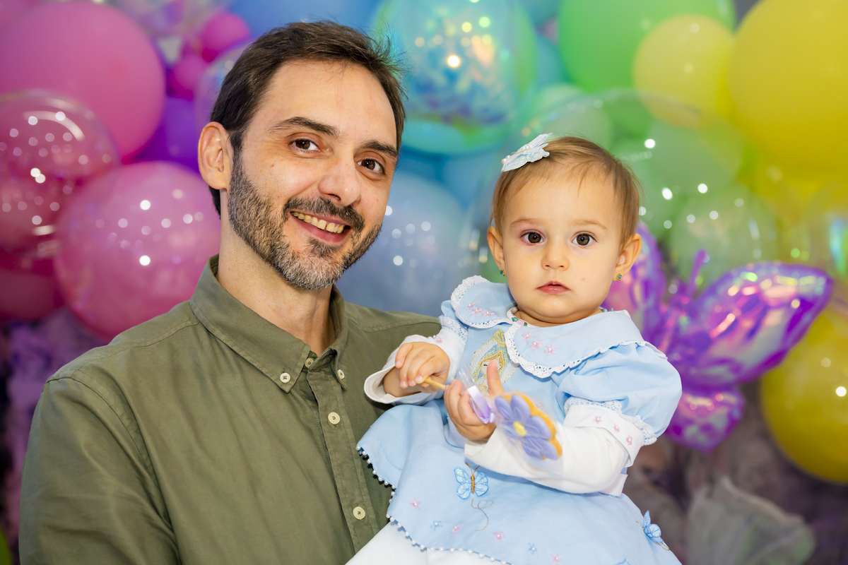 Pai segura a filha aniversariante no colo em frente ao painel colorido de balões, compondo registro afetivo da festa infantil em Alphaville.
