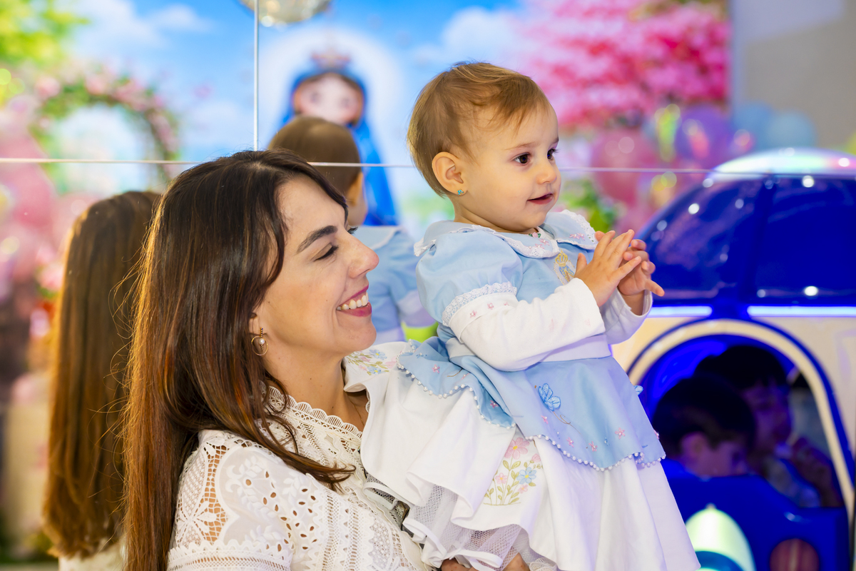Madrinha carrega a aniversariante no colo próxima a um carrinho de brinquedo, com painel florido ao fundo no salão temático em Alphaville.
