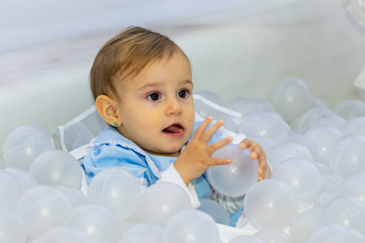 Aniversariante de vestido azul explora a piscina de bolinhas com expressão curiosa, cena descontraída da festa infantil em Alphaville.