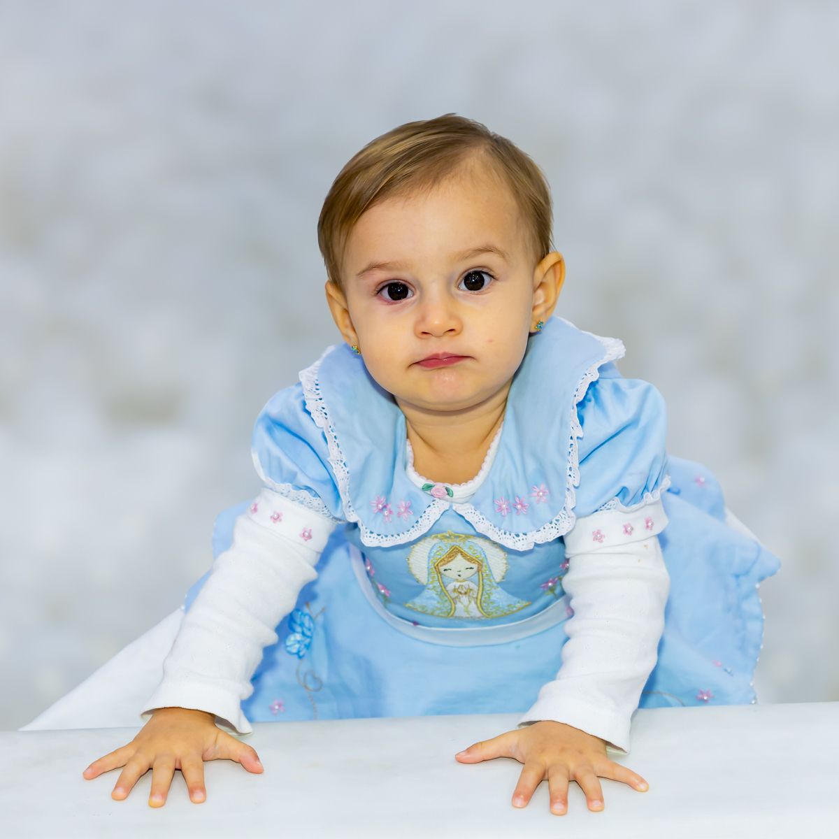 Retrato de estúdio da aniversariante com vestido azul bordado, apoiada em superfície branca, destacando o traje especial da comemoração.