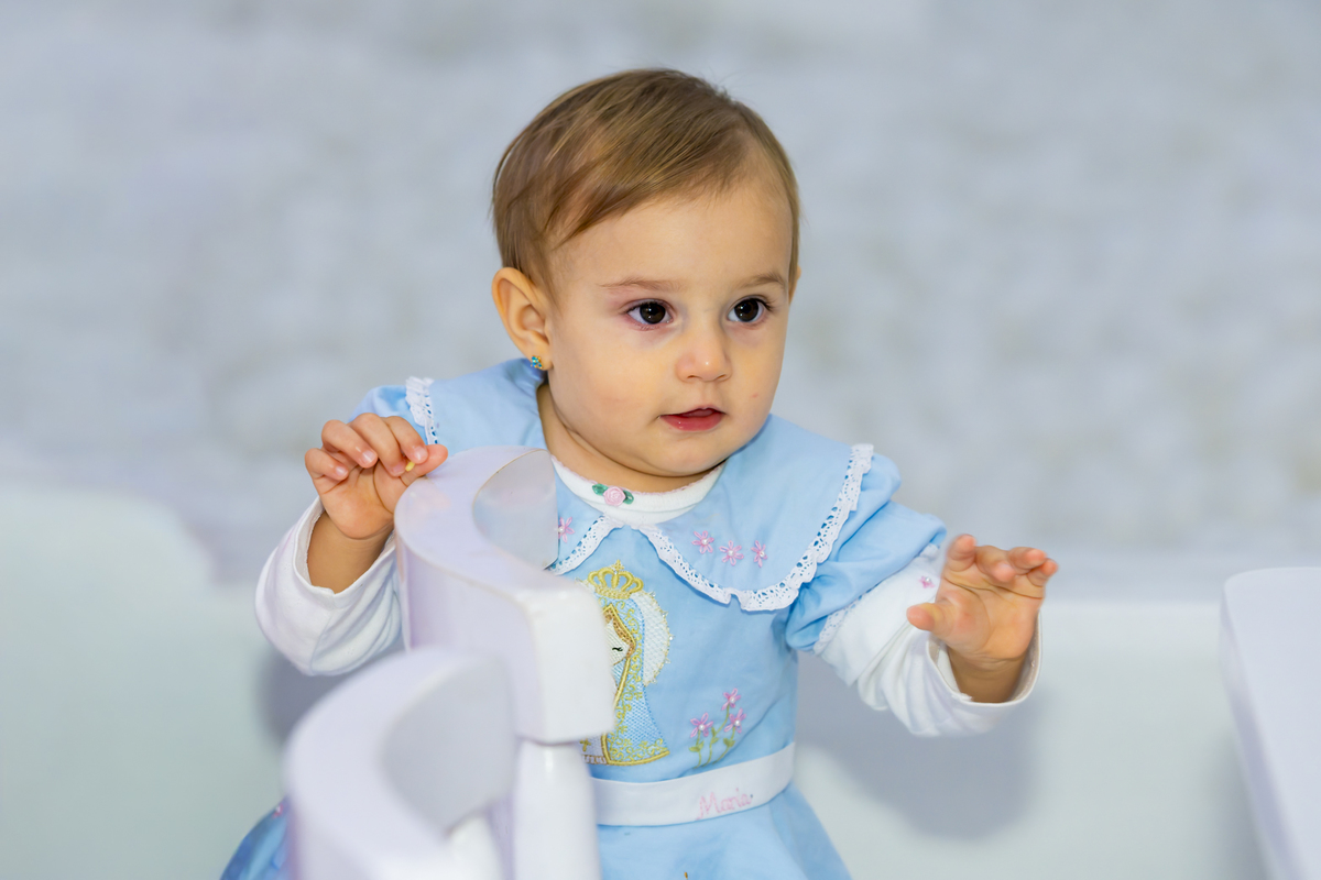 Bebê aniversariante observa curiosa ao lado de um brinquedo branco, em ensaio fotográfico realizado no espaço Lá Vem o Sol - Lua, Alphaville.