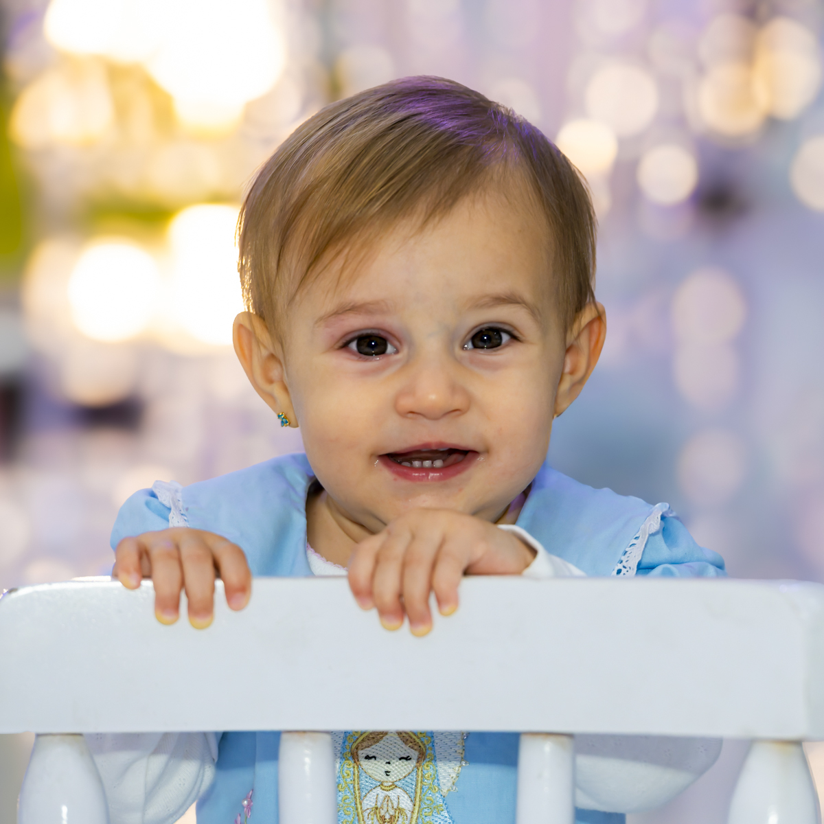 Aniversariante sorri apoiada em cadeirinha branca durante festa de aniversário no Lá Vem o Sol Lua Alphaville. Luzes de fundo em bokê dourado destacam o vestido azul com detalhes bordados.