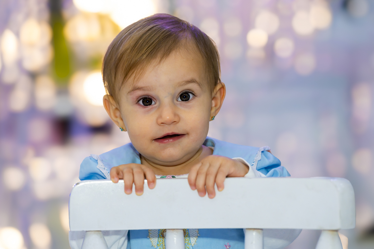 Bebê observa atentamente a câmera durante comemoração de aniversário em Alphaville. Fundo desfocado com luzes suaves realça a expressão curiosa da pequena.