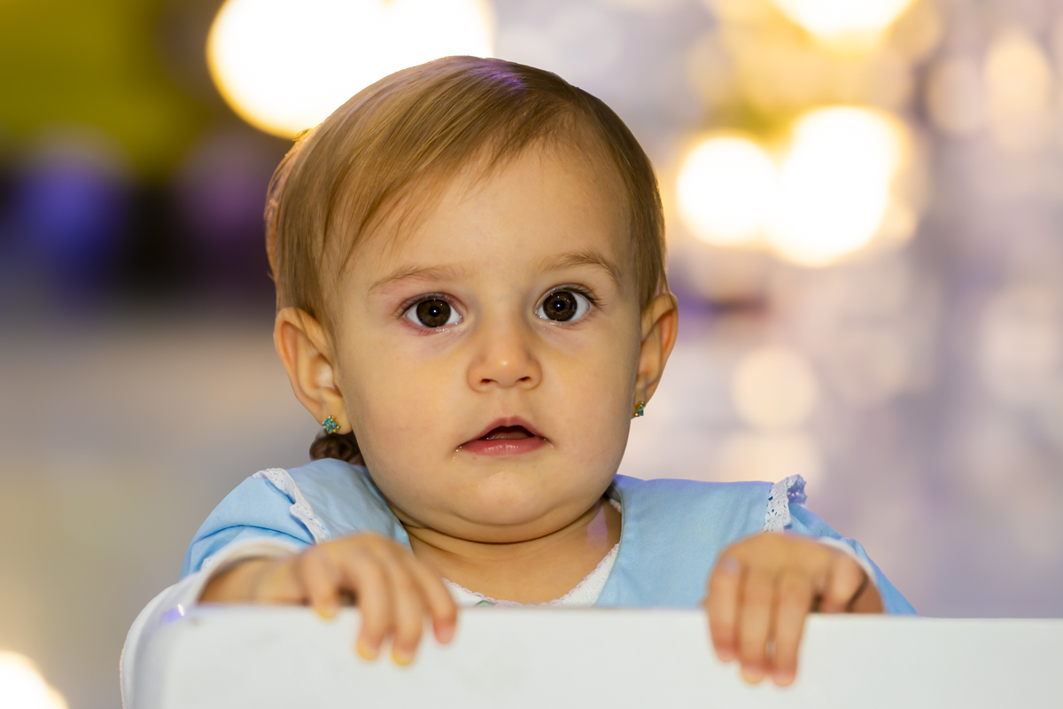 Close da aniversariante apoiada na cadeira branca em ambiente iluminado de festa infantil em Alphaville. Expressão serena reforça a atmosfera acolhedora da celebração.