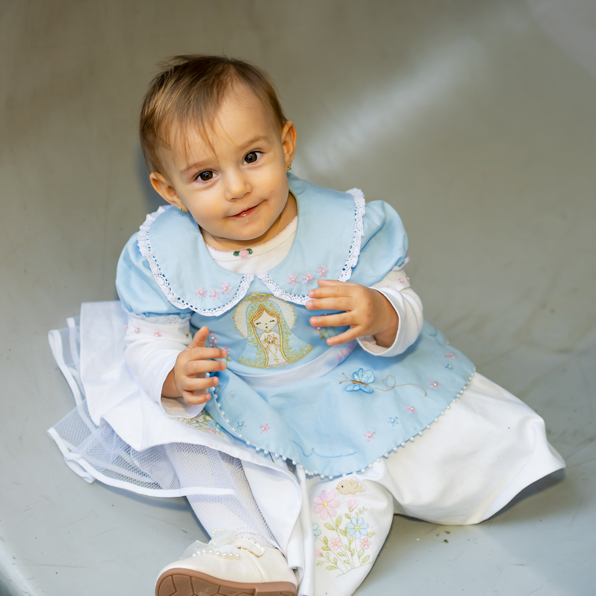 Bebê aniversariante sorridente brinca sentada durante sessão de fotos de aniversário no Lá Vem o Sol Lua. Vestido bordado azul e branco reflete o tema da celebração.