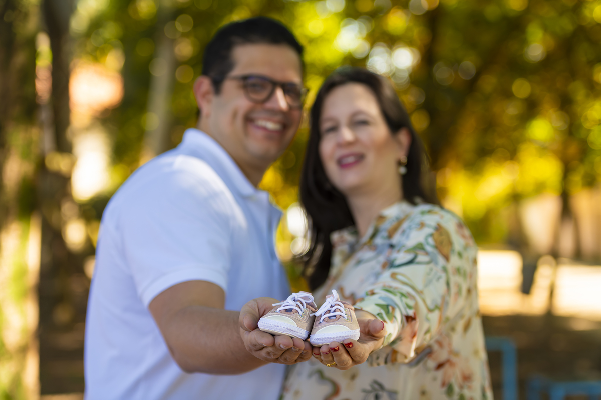 Casal sorridente segura sapatinhos de bebê em ensaio ao ar livre na praça, celebrando a espera do novo integrante da família.