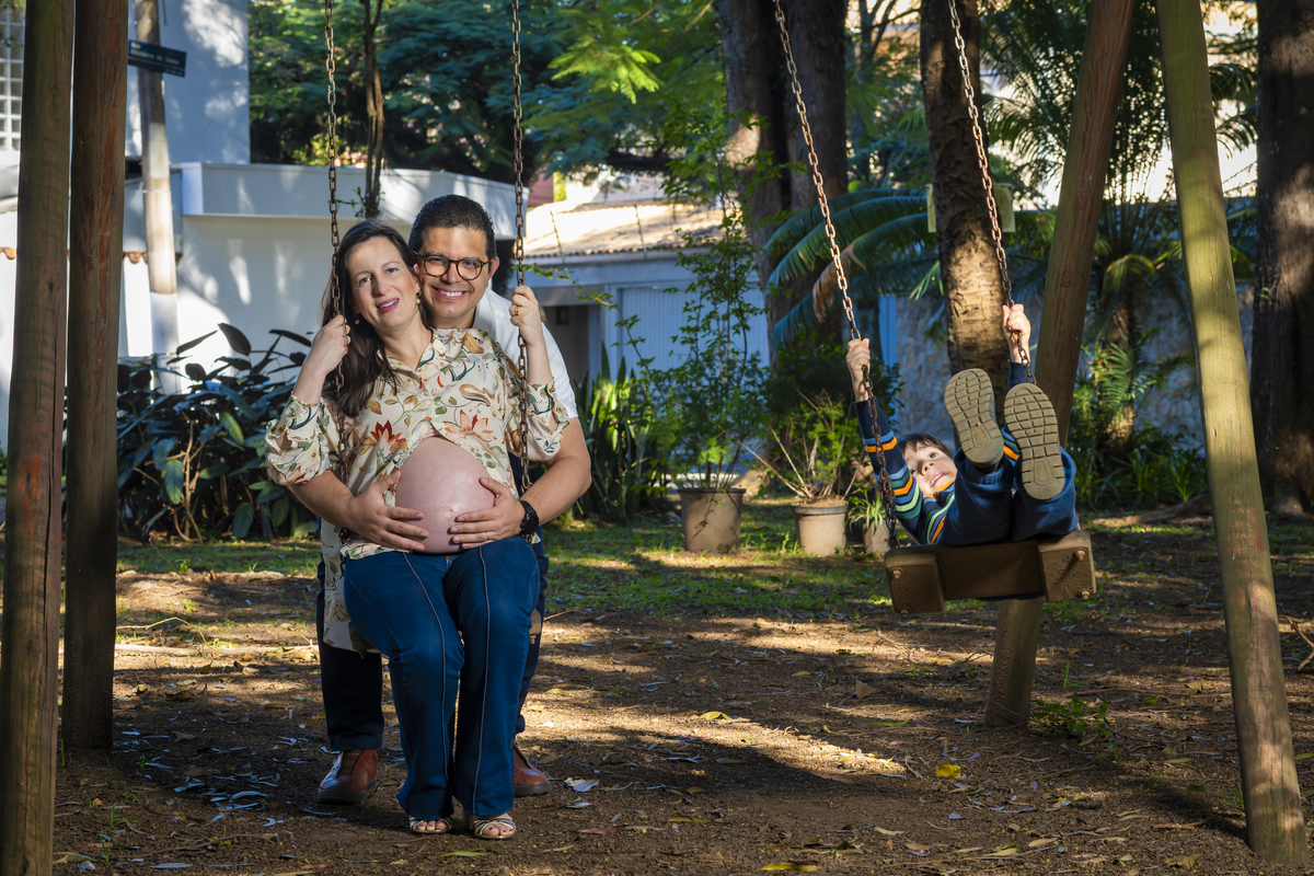 Casal se abraça em balanço de madeira na praça enquanto o filho brinca ao fundo, cena espontânea de ensaio de gestante em família.
