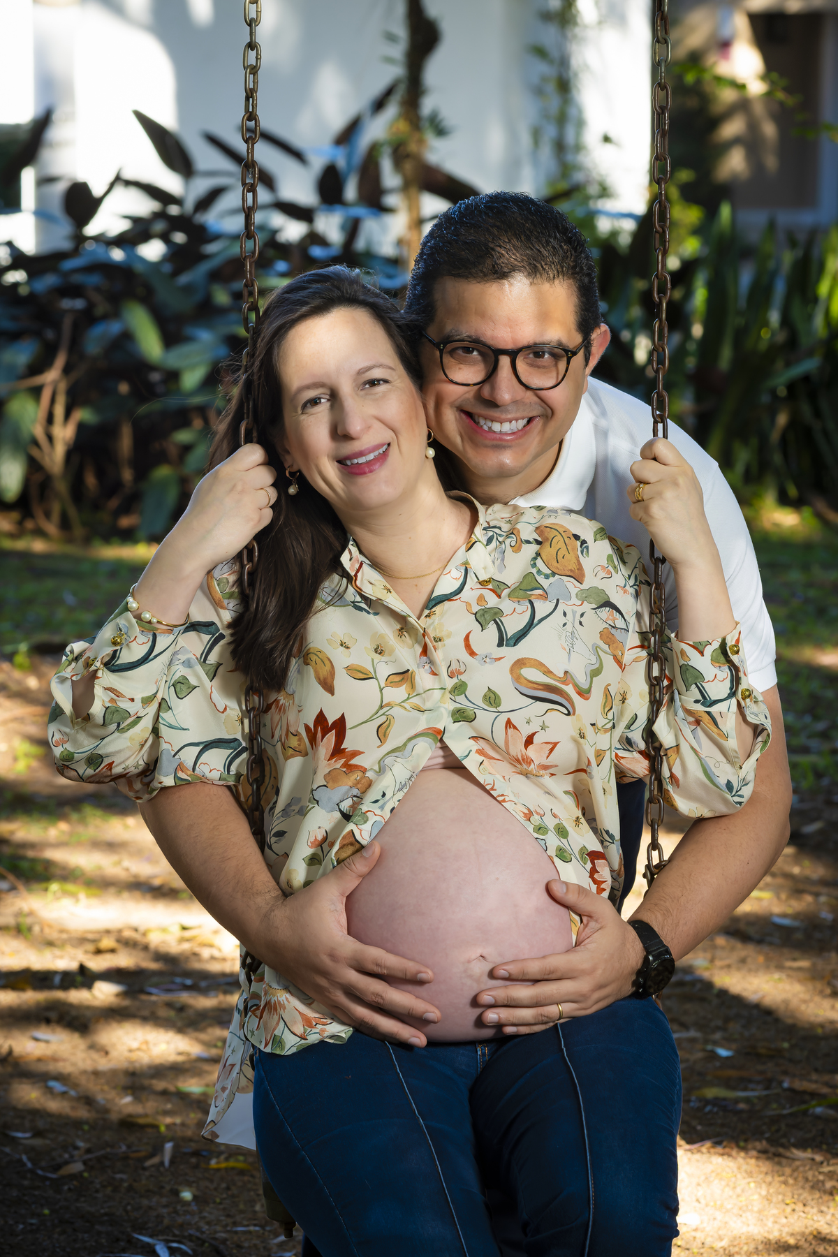 Close do casal sorridente no balanço, com as mãos na barriga da gestante, retratando ternura durante ensaio ao ar livre em São Paulo.