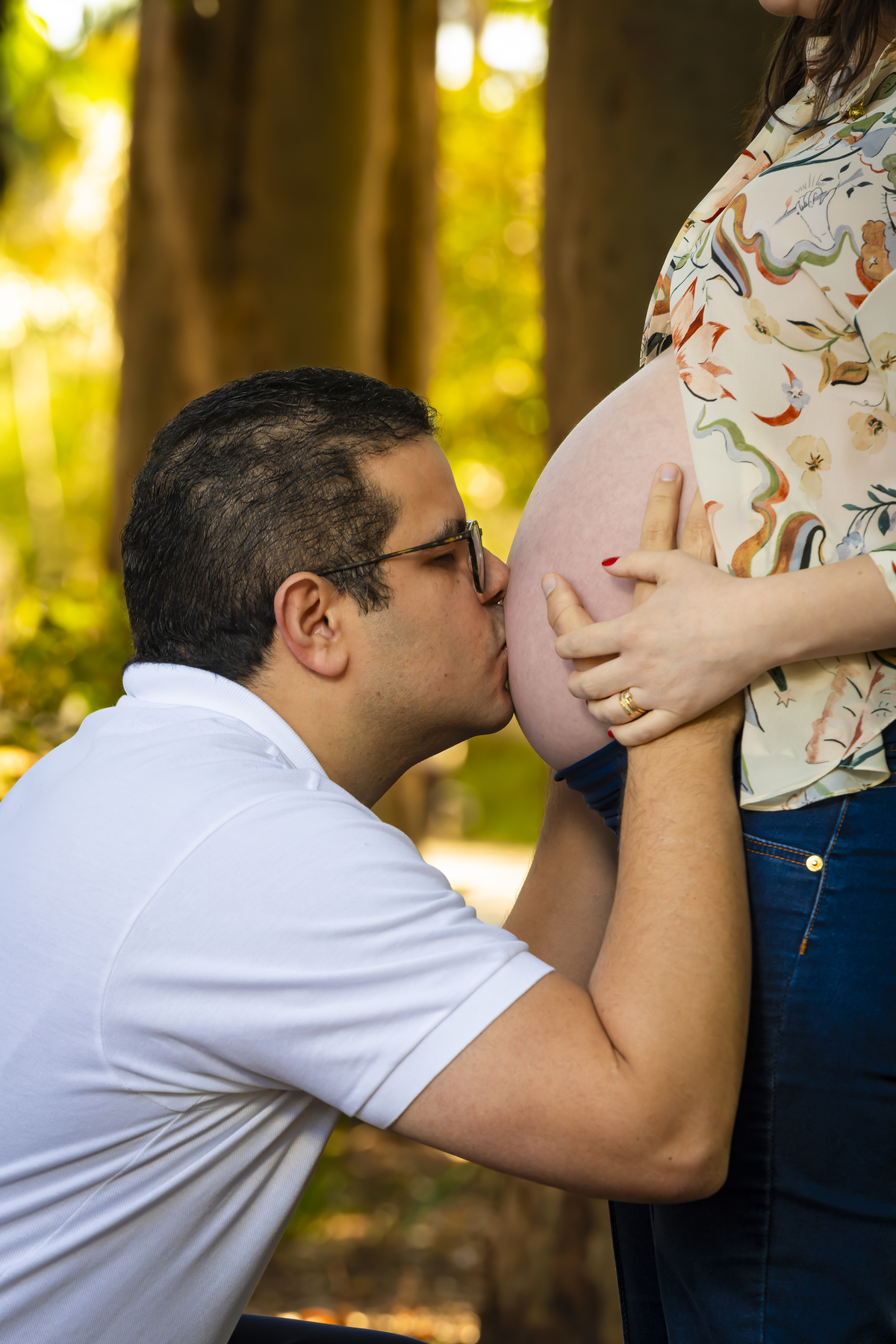 Momento delicado em que o pai beija a barriga da gestante entre as árvores, cena emocionante de ensaio fotográfico em família.