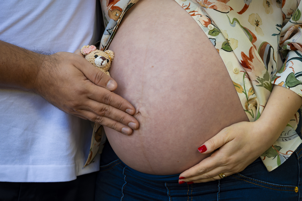 Detalhe das mãos do casal segurando um ursinho de crochê sobre a barriga da gestante, simbolizando a espera do bebê.