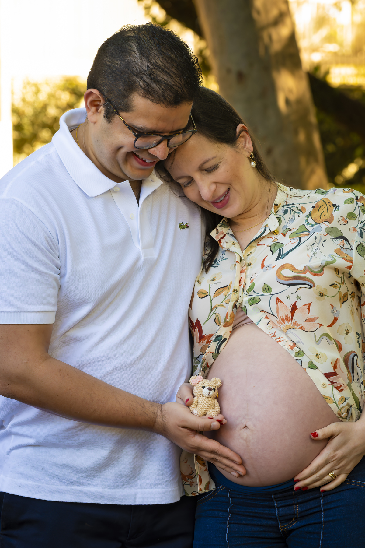 Casal sorri em momento íntimo, com as mãos unidas sobre a barriga, cercado pela luz dourada da tarde na praça  Conde Barcelos em São Paulo,