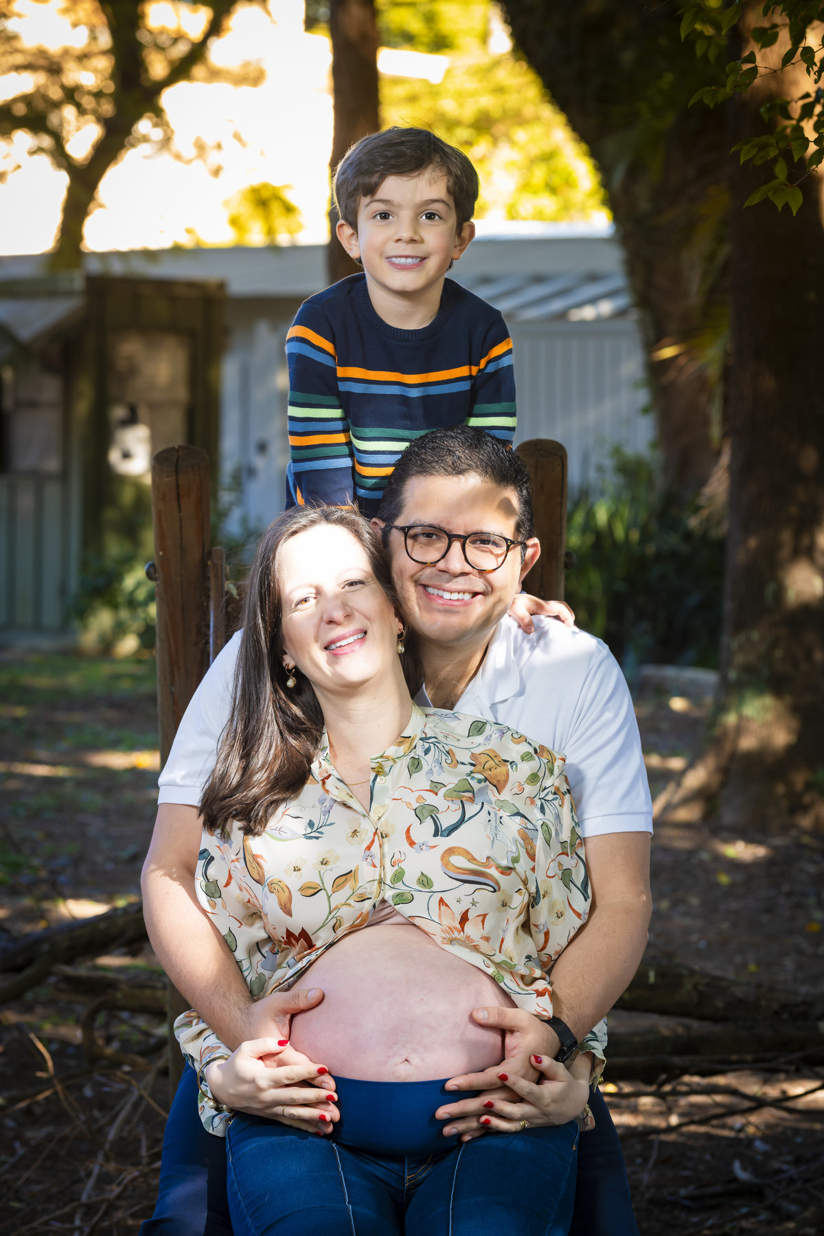 Retrato em close da família sorrindo, com destaque para a barriga da gestante, em ambiente arborizado da praça Conde Barcelos em São Paulo.