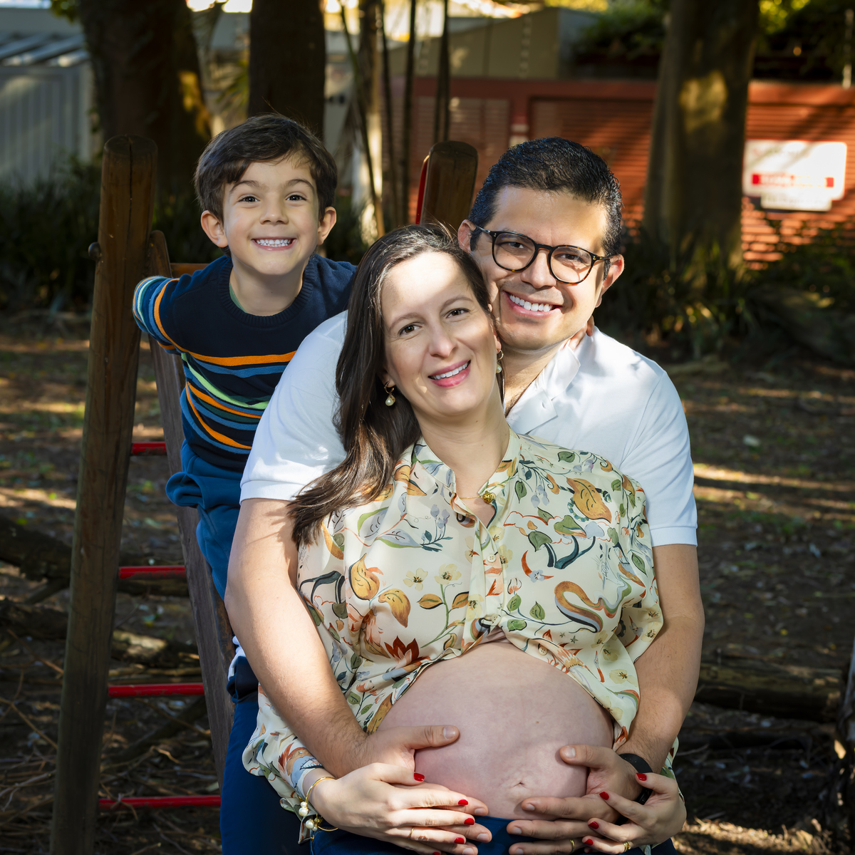 Família posa unida no playground, com o filho brincando na estrutura de madeira, retratando alegria e conexão familiar.