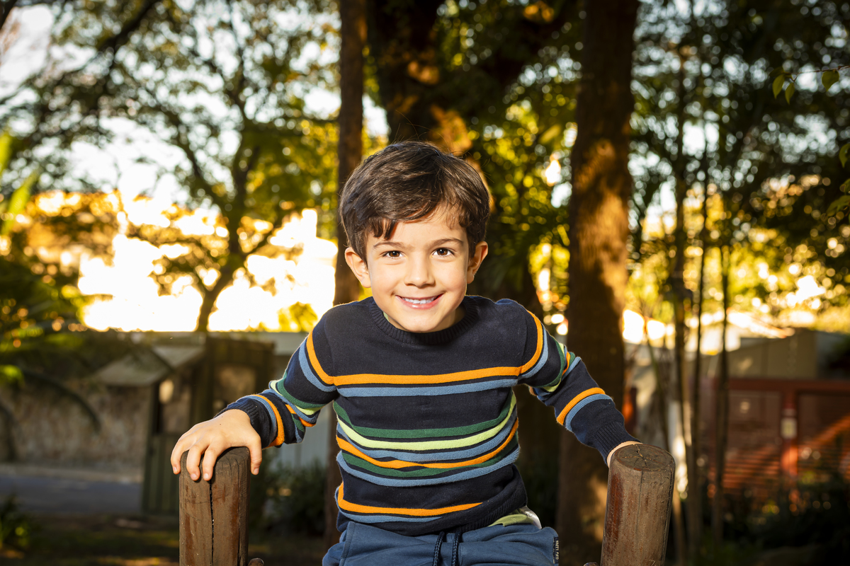 Retrato individual do filho sorridente apoiado em estrutura de madeira, com luz dourada do entardecer ao fundo.
