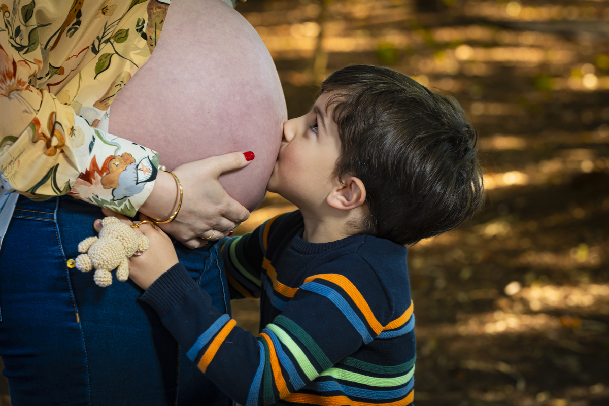 Irmão mais velho beija a barriga de grávida em ensaio family na Praça Conde Barcelos em São Paulo, segurando ursinho de crochê. Momento afetuoso que celebra a chegada do novo bebê.