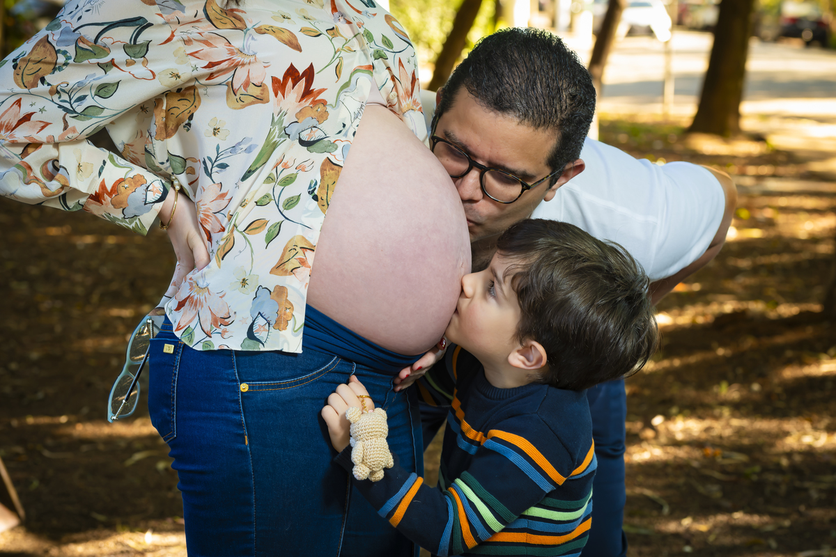 Close do pai e do irmão mais velho beijando a barriga da mamãe grávida, cercados pela vegetação da praça Conde Barcelos em São Paulo. Detalhe do ursinho de crochê reforça o clima aconchegante do ensaio.