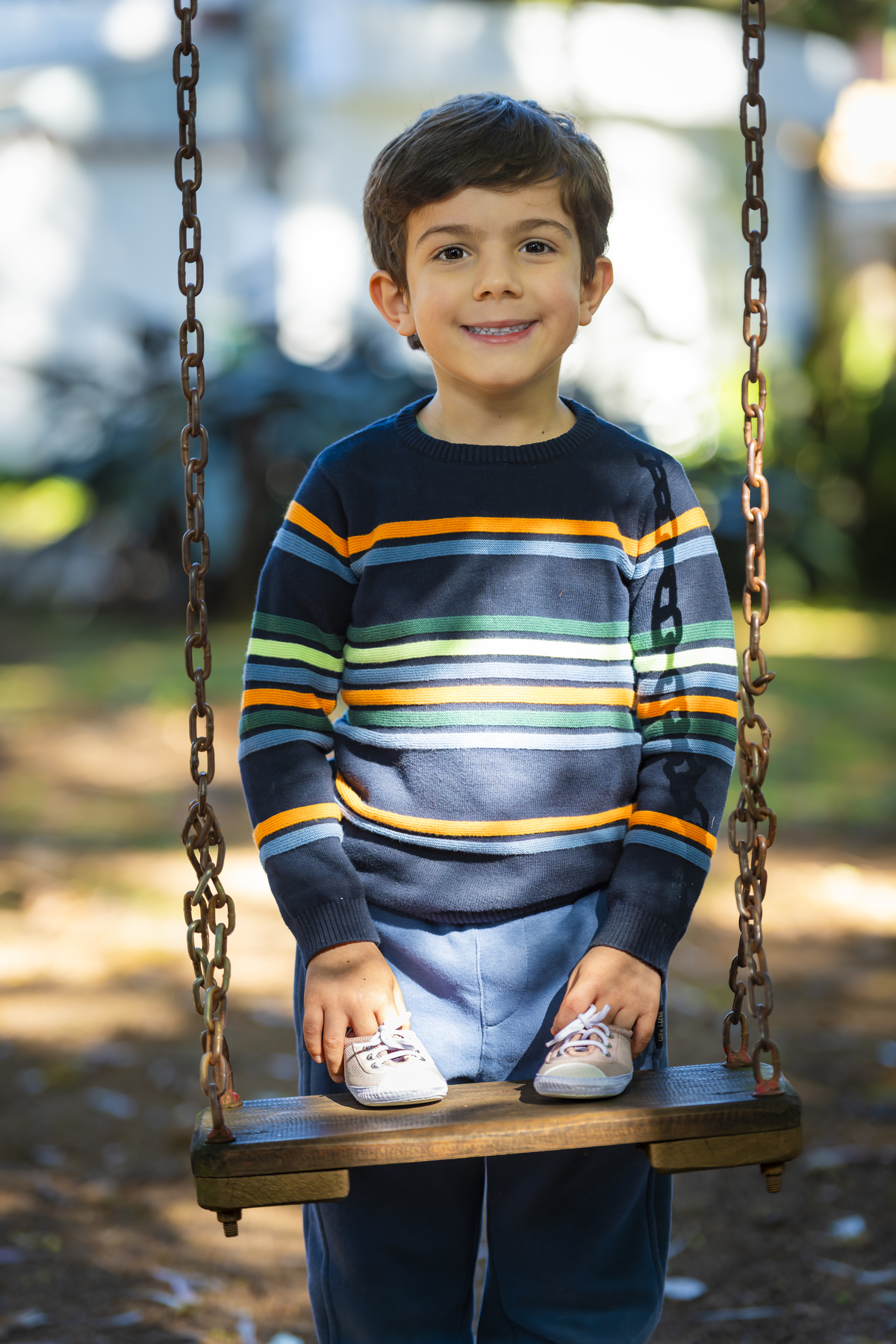 Menino sorridente posa em cima de balanço de madeira segurando os tenistas do futuro irmãozinho, em praça de São Paulo. Retrato espontâneo que expressa alegria durante o ensaio family.