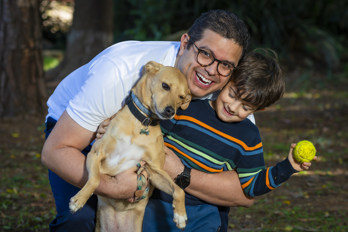 Pai sorri ao abraçar o filho e o cachorro da família, momento descontraído em meio à vegetação da praça. Cena espontânea que reforça o clima leve do ensaio ao ar livre em São Paulo.
