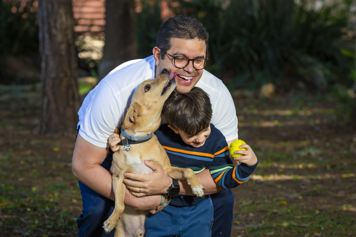 Cachorro lambe o rosto do pai enquanto ele abraça o filho, gerando risadas durante o ensaio family na praça. Registro divertido e autêntico da interação entre pet e família.