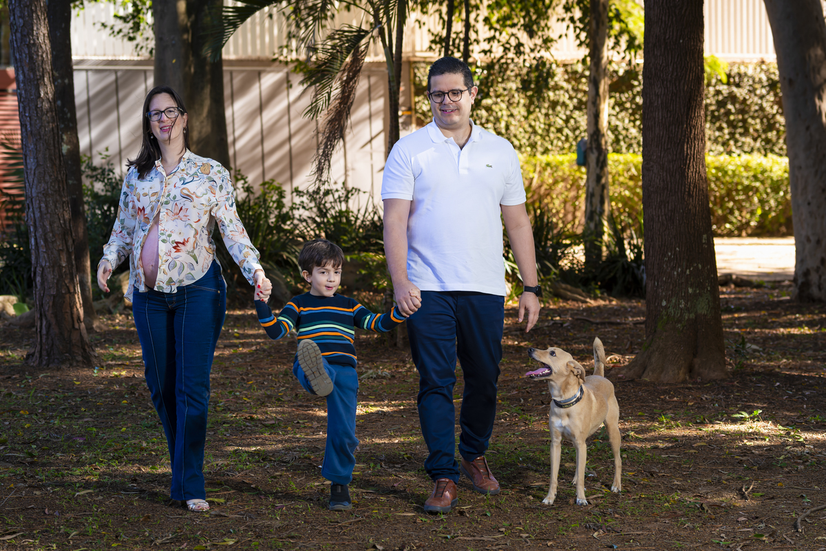 Família caminha de mãos dadas com o cachorro por entre as árvores da praça em São Paulo. Cena natural que retrata o passeio em família durante o ensaio de espera do bebê.