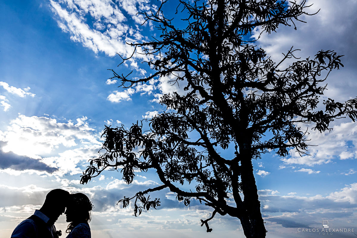 contra luz com o céu azul de fundo com uma árvore junto ao casal fotografo de casamento três corações mg carlos alexandre foto casamento em são thomé das letras sul de minas