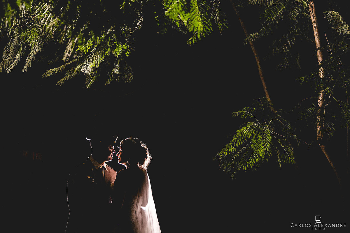 fotografia de contra luz de casal de noivos, no meio de galhos e folhas verdes, noivo militar com terno característico, fotografado por carlos alexandre, da cidade de 3 corações, e sul de minas