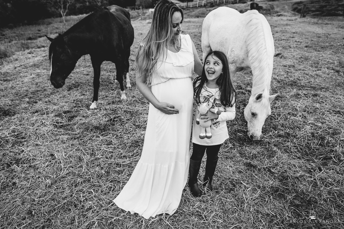 tia e sobrinha brincam ao falar da bebê que está na barriga, com os cavalos ao fundo, no ensaio de gestante em três corações carlos alexandre foto