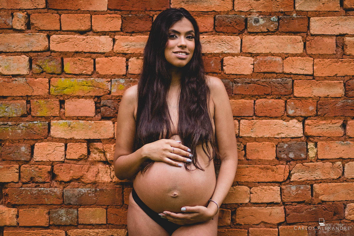 menina com os seios à mostra, com seus belos cabelos longos e pretos tapando os seios, enquanto segura e acaricia sua barriga, fotógrafo de gestante em três corações mg carlos alexandre fotos em fazenda cachoeira